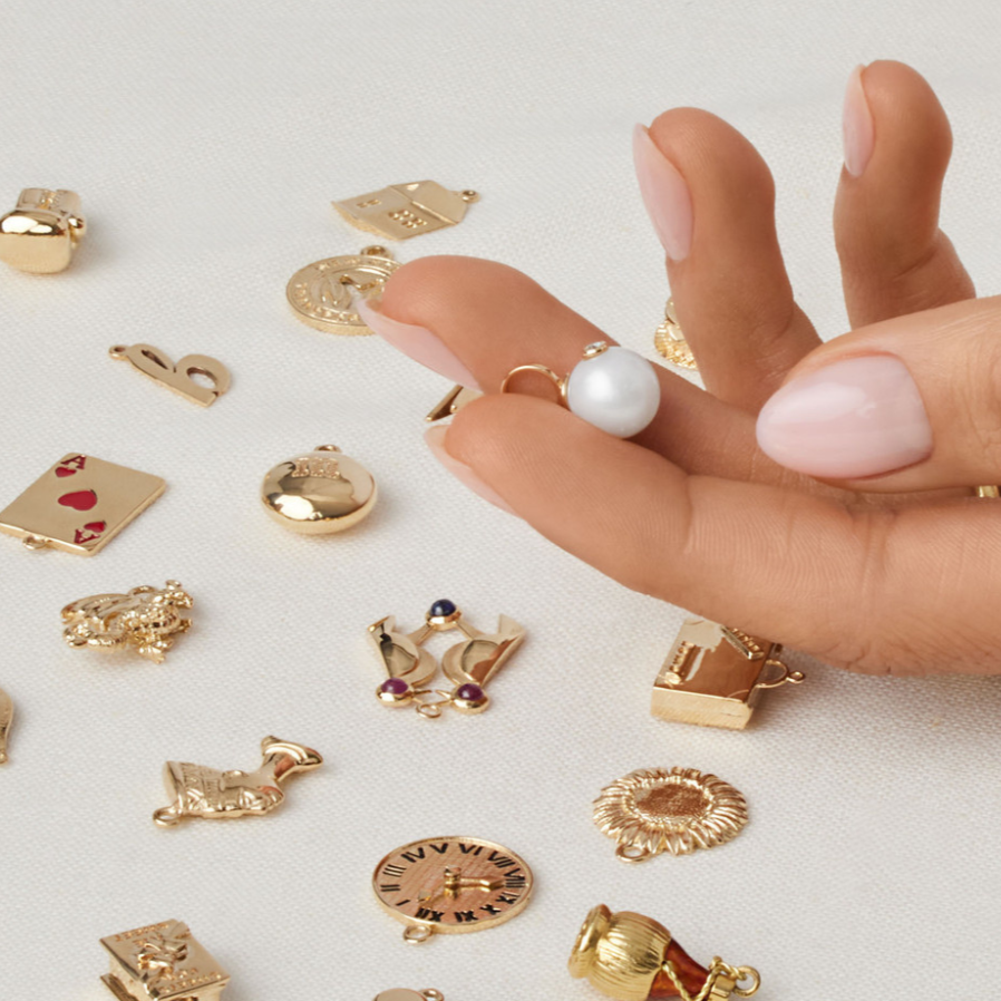 Gold charms on a white surface with a hand wearing a pearl ring.